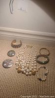 Photo shows a full lot of 3 bracelets, 2 brooches, and a faux pearl necklace arranged on carpet.