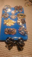 Ten pieces of costume jewelry displayed on a blue board, including brooches with floral and abstract designs in gold-tone metal with colored stones, and a black bow-shaped brooch.