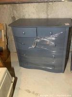 Front view of blue painted wood dresser with five drawers, wrapped in plastic on center drawers.