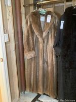 Full view of the long muskrat fur coat hanging on a rack next to a black fur coat.