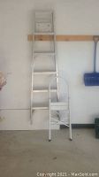 View of an aluminum ladder standing upright against a white wall, with a smaller white folding step stool in front.