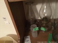 Two oil lamps on a shelf with visible height labels '4.5' and '12' inches on green stickers. The lamp on the right has a brass burner and clear glass chimney.