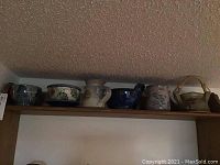Full view of seven pottery pieces displayed on a wooden shelf including bowls, jugs, plate, and tea pot.
