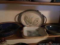Four pottery items displayed on shelf: deep blue bowl with moose design, oval tray with handles and leaf design, rectangular tray with green leaf pattern, and large round pottery item with faint engraved pattern.