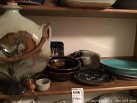 Photo of pottery items on wooden shelf, including large beige and brown container with spigot, multiple plates and bowls in various colors and styles.