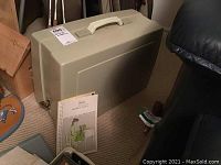 Closed beige carrying case with handle, instruction booklet standing beside it on carpet, lower corner of wooden table and part of black chair visible.