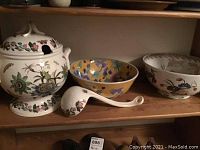 Soup tureen with lid and ladle on wooden shelf alongside two decorative bowls.