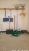 Wide view of all yard tools including shovels, rakes, brooms, garden hose, and extension cord arranged standing against wall.