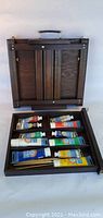 Closed wooden foldable art set case with handle, opened to show trays inside with paint tubes.