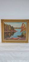 Full view of the signed oil painting showing autumn lakeside landscape with trees and rocks, framed in decorative gold frame.