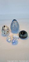 Front view of five art glass pieces on plain background showing multicoloured Boda egg, blue egg, black round paperweight, and two small white eggs with blue swirls