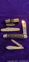 Photo showing miniature Hohner harmonica with advertising caps 'HOH NER Made in Germany', metal tag '1933-1934', and two folding knives including one Imperial folding knife with textured handle.