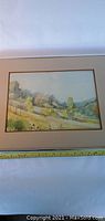 Framed watercolor artwork showing landscape scene with trees, hills, and field. View includes measuring tape for size reference.