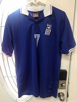 Front view of an authentic Greece national soccer team jersey in blue with white collar and trim, number 7 and Greek flag emblem on chest.