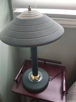 Side view of green metal lamp showing round conical shade with rings, brass accent at base, and green cylindrical stand.