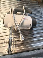 Side view of white Sunbeam hand mixer with black handle and two metal beaters inserted, cord wrapped around handle.
