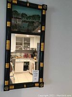 Full view of antique mirror showing black and gold frame with a reflective lower section and decorative painted panel at the top depicting a countryside scene.