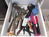Top-down view of a drawer containing assorted cooking utensils including wooden-handled spoons, metal skimmer, ladle, scissors, plastic knives, and measuring cups and spoons.