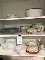Side view of shelf showing stacked dishes including decorative bowls and plates, white square bowls, and casserole dish.