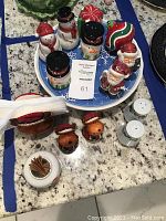 Image showing assorted holiday-themed salt and pepper shakers on a white and blue round plate, including snowmen, Santa Claus, and reindeer figures alongside small white salt and pepper shakers, a ceramic turkey napkin holder and a toothpick holder.