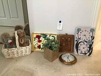 Wide shot showing white wicker basket holding four stuffed bears, framed wood carving, faux plant, round clock, floral clock/thermometer on floor