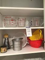 Three Pyrex glass measuring cups on shelf with red measurement markings, two plastic colanders (red and yellow), metal sifter, cone-shaped cheese grater with wooden handle, and round metal strainer.