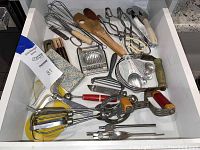 Photo of a drawer filled with assorted vintage kitchen utensils including a hand mixer, egg slicer, peelers, whisks, tongs, can openers and a pizza cutter.
