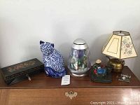 Photo showing all 6 items: black painted box with floral design and key, blue and white ceramic cat figurine, simulated aquarium light, brass lamp with floral shade, painted heavy metal figurine, and vintage miniature camera.