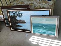 Three framed paintings leaning against a table, showing two landscape/seascape artworks with detailed frames.
