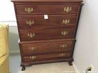 Front view of wooden chest of drawers showing three top drawers and two lower drawers with brass handles and bracket feet.