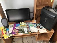 Dell computer monitor, speakers, assortment of puzzle and crossword books, paper shredder, and various papers and cards on a wood desk with wood shelving behind.
