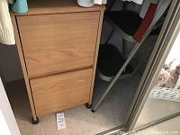 Photo of two drawer filing cabinet with wood grain finish on black wheels next to green metal frame 2-step folding ladder.
