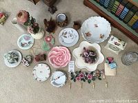 Photo showing a collection of assorted ceramic and porcelain decorative items including floral plates, vases, a hook rack with pink rose hooks, and a large pink rose plate.
