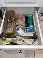 Photo of open drawer with assorted household tools and hardware items including hammers, paint brushes, flashlights, and miscellaneous nails and screws