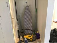 Yellow vacuum cleaner with hose, striped ironing board leaning against a door, Swiffer duster with green handle is partially visible.