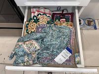 Top view of a drawer showing folded kitchen textiles including floral and patterned oven mitts, pot holders, and towels.