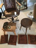 Photo of brown leather swivel chair, rectangular step stool, round vintage adjustable height stool, brass door knocker, and five carved wood wall art plaques.