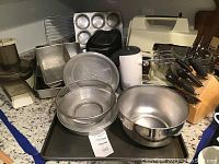 Shows general overview of the lot items including mixing bowls, bakeware, appliances, sieve, knife block with knives.