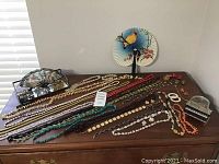 Full display of assorted necklaces arranged in rows on a wooden surface with a glass jewelry box and two decorative handheld fans in the background.