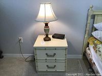 Straight-on view of white nightstand with table lamp and alarm clock on top next to bed