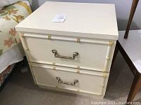 Photo of a white painted nightstand with two drawers featuring decorative bamboo style trim and brass or gold-tone pull handles.