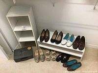 Two white storage shelves of different sizes, assorted women's shoes arranged on and near one shelf, and a black fire resistant lockbox with a key on top, all placed against a white wall.