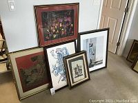 Photo showing five framed prints leaning against a wall and door on carpeted floor in a home setting.