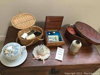 Photo showing wooden chest with key, teacup and saucer pin cushion, hat pin cushion, small woven basket containing thread and scissors, small wooden box with sewing supplies, and oval wooden basket with sewing items.