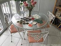 Full view of round wrought iron table with clear glass top, 4 wrought iron chairs with pink upholstered cushions around table, vintage coasters and floral decor on table