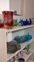 Shelf with red glass vase, green bowl, blue glass trinkets and bottles, grey glass bowl with fruit patterns, and blue lidded glass dish.