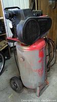 Side view of vertical air compressor with red tank, black motor on top, wheels and handle at base.