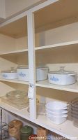 Shelf with multiple CorningWare covered casserole dishes with blue floral pattern, glass lids, along with Pyrex clear glass dishes and plastic serving pans