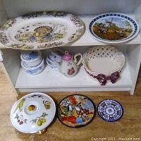 Wide view of two shelves holding assorted used dishware including the turkey platter, painted plate, ceramic jars, teapot, small basket, and round plates on the floor.