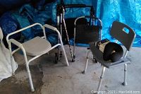 Black folding walker with wheels, two patterned canes, white metal toilet top with padded seat and armrests, gray shower seat with backrest holding additional accessories. Blue tarp backdrop.
